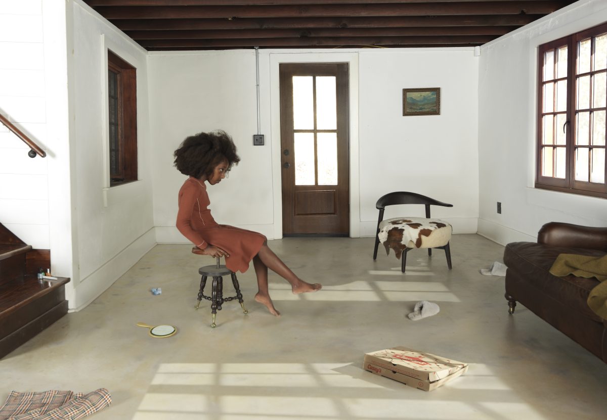 A woman in a rust-colored dress sits on a metal stool in a sparse, minimalist interior with white walls and concrete flooring. Sunlight streams through windows and an open doorway, casting geometric shadows across the floor scattered with domestic objects including plates, books, and textiles, while furniture and a staircase are visible at the room's edges.