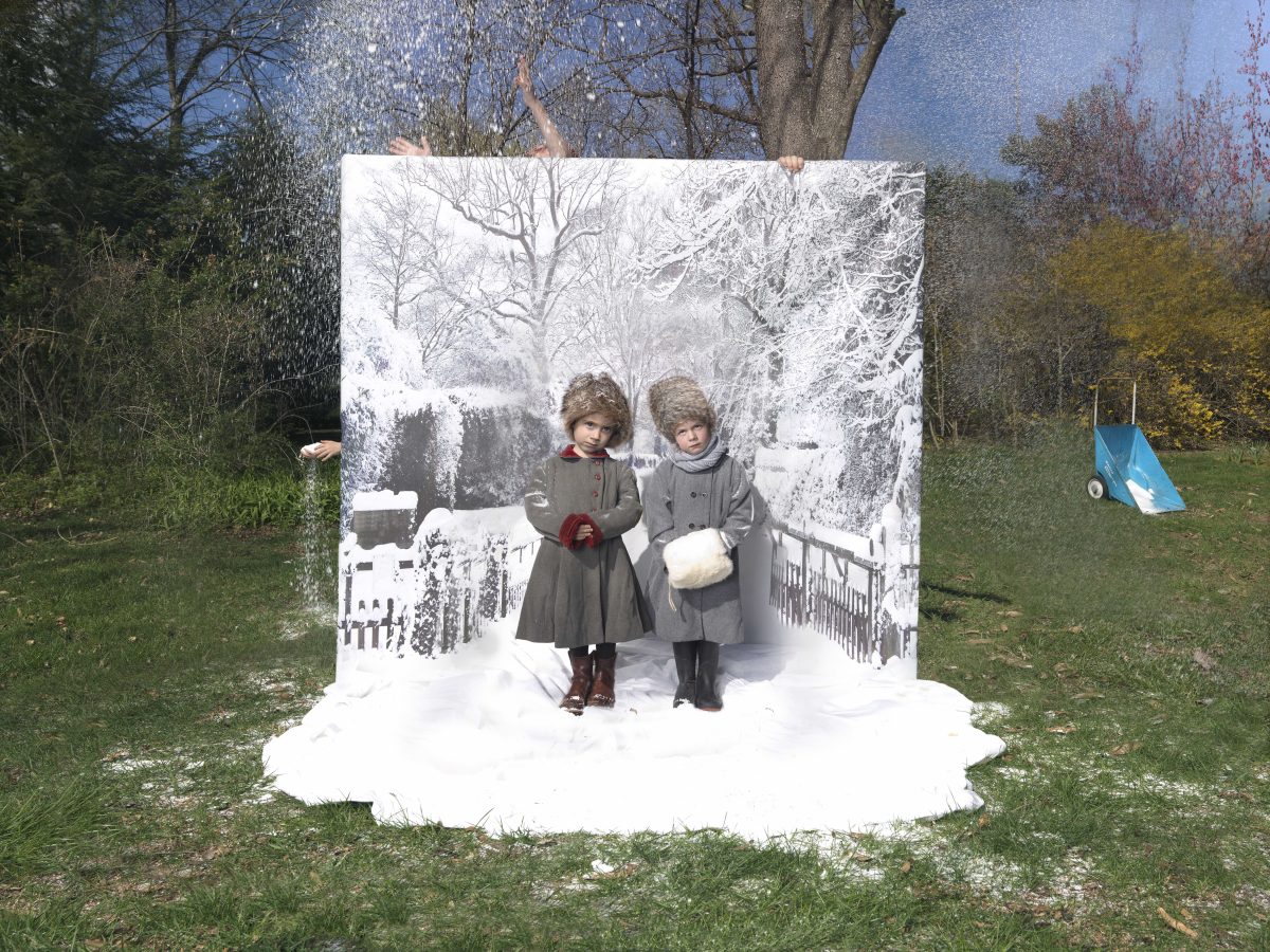 Two young children in winter coats and knit hats stand on artificial snow in front of a large white panel depicting a snowy landscape with bare trees and a fence. The panel is positioned in a green outdoor setting with bare trees visible above it, creating a striking contrast between the constructed winter scene and the actual temperate surroundings.
