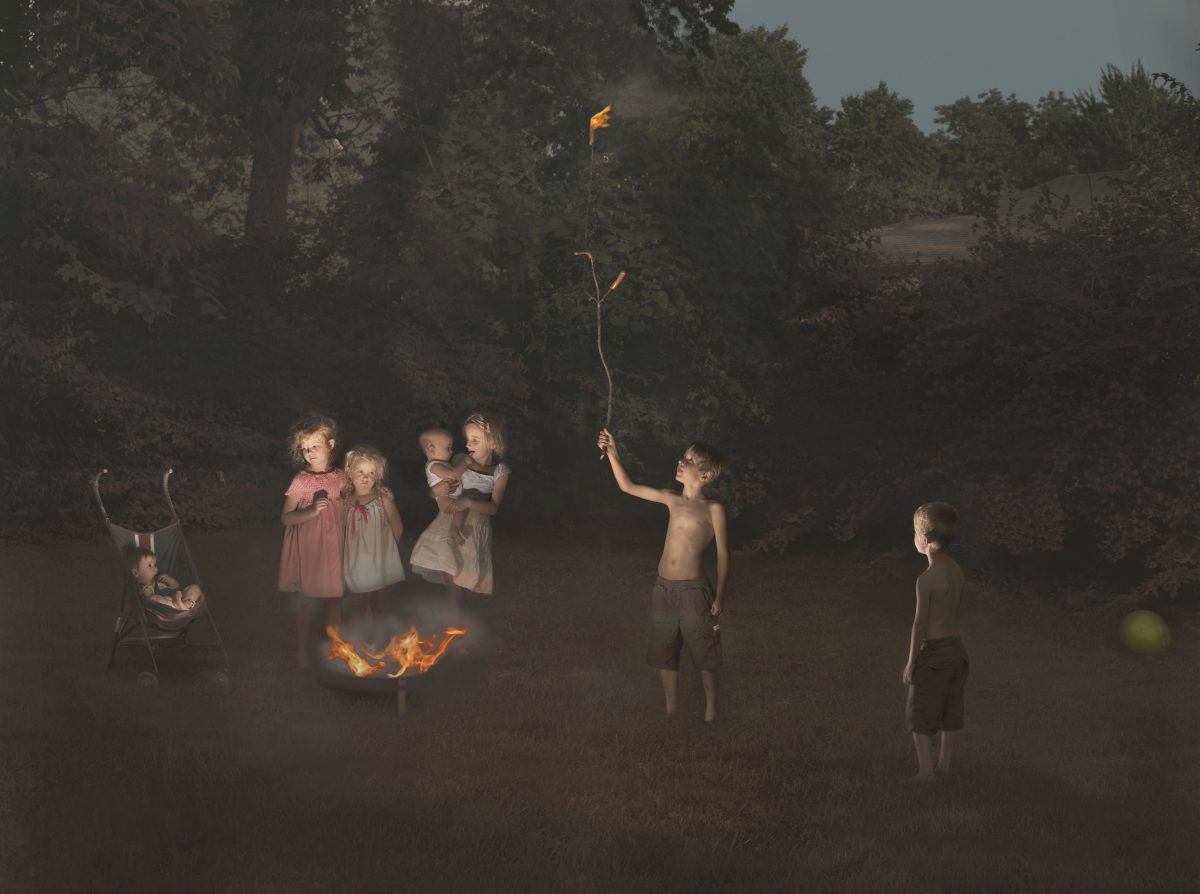 Children gathered in a misty forest clearing around a campfire, with one child holding aloft a flaming torch against dark trees. The scene is rendered in muted, atmospheric tones with dramatic lighting that isolates the figures from the shadowy woodland background.