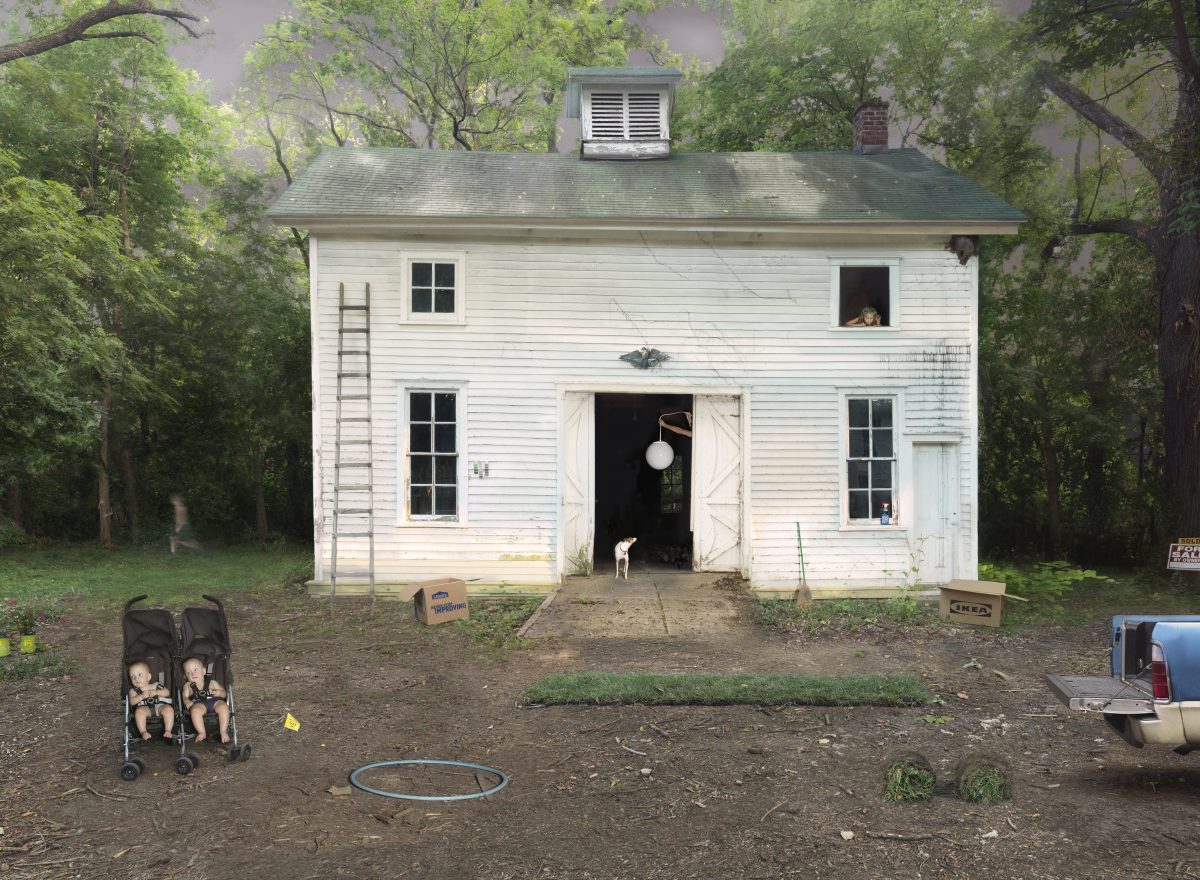 A white wooden house surrounded by dense green trees serves as the backdrop for a domestic scene. In the yard, a double stroller with children sits in the foreground, while various household items, toys, and construction materials are scattered around the property, including an IKEA box and what appears to be items in the process of being moved or organized.