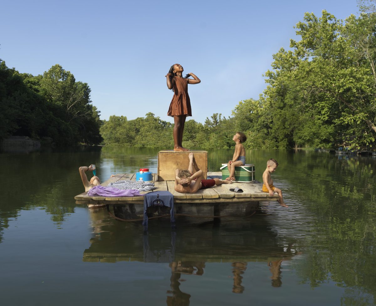 A bronze sculptural figure of a girl stands on a wooden platform in the center of a calm river, shading her eyes as she gazes into the distance. Around her, several children in various states of repose occupy the platform and its edges—one lying down, others sitting or dangling their feet in the water—surrounded by coolers, supplies, and domestic objects, creating a surreal tableau of leisure amid lush green trees.