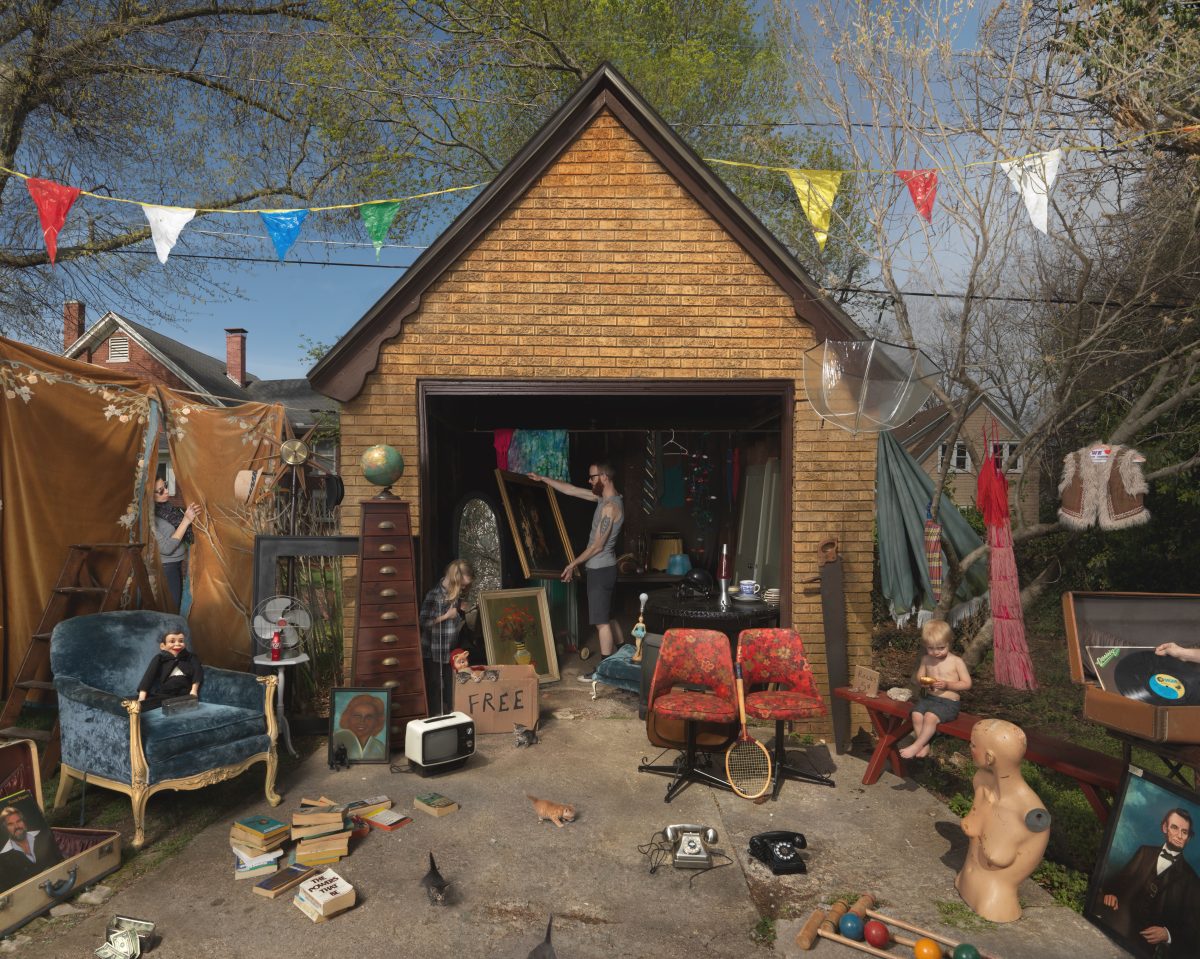A brick garage with open doors displays an eclectic collection of secondhand items arranged in the driveway and surrounding area, including furniture, artwork, books, vintage telephones, mannequins, and clothing on clotheslines. Colorful bunting flags stretch overhead while several figures, including children and adults, interact among the goods, creating a vibrant scene of domestic clutter and commerce.