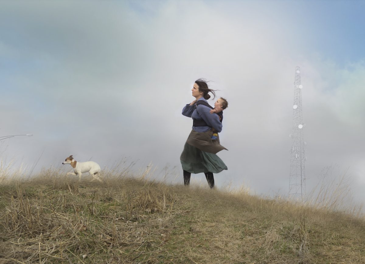 A woman holding a young child stands on a grassy, windswept hilltop, gazing into the distance. A small white dog walks nearby, while a tall communication tower looms through the misty fog in the background under an overcast sky.