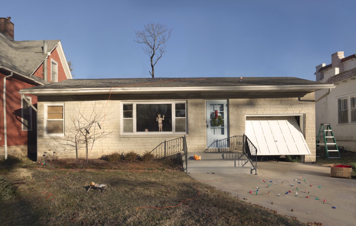 A suburban single-story house with cream-colored brick exterior and large front window displaying a figure in silhouette. The concrete driveway is scattered with colorful toys, while bare branches and holiday decorations accent the modest front yard and porch area. Neighboring residential buildings frame the composition under clear blue sky.