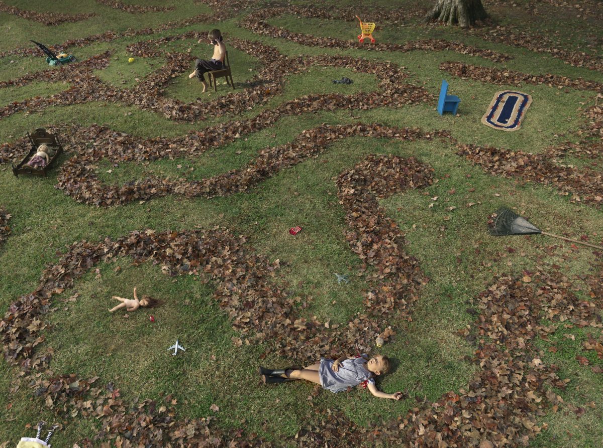 Aerial view of a suburban lawn scattered with fallen leaves and children's toys, featuring two shirtless figures lying on the grass among the leaf piles and a seated child in the upper portion of the composition, rendered in saturated color photography.