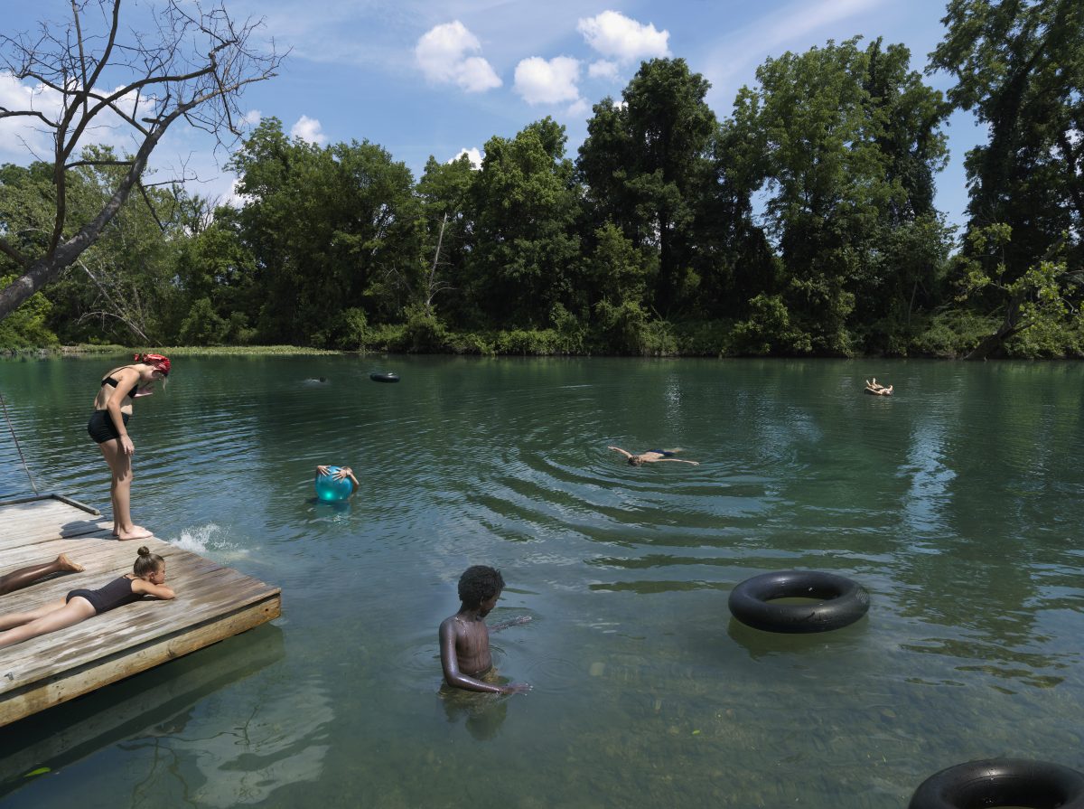 A group of people swim and play in a clear green river lined with dense trees, with wooden docks in the foreground where others rest and prepare to enter the water. Black inner tubes float on the water's surface as swimmers of various ages enjoy the natural swimming hole on a sunny day.