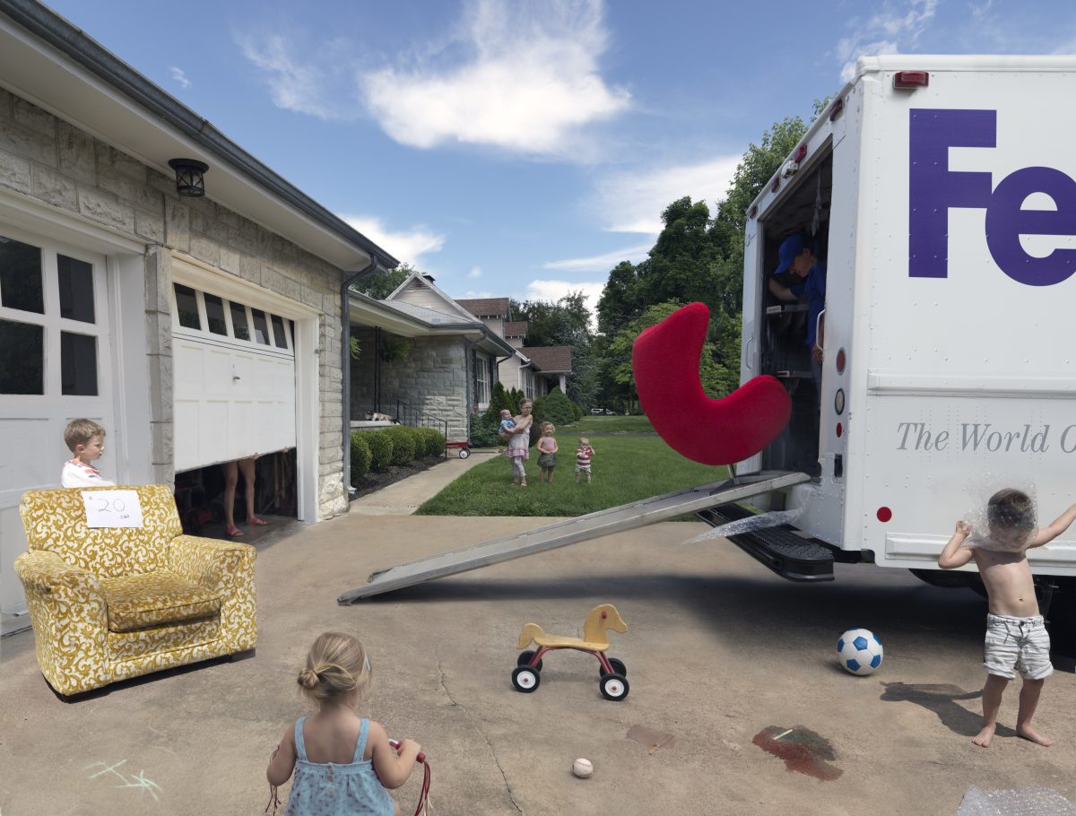 A suburban driveway scene with children playing among oversized objects: a giant yellow patterned armchair on the left, a large red curved form mounted on a delivery truck on the right, and scattered toys including a wooden duck-shaped rider and soccer ball. Residential houses and green lawn visible in the background under a blue sky.