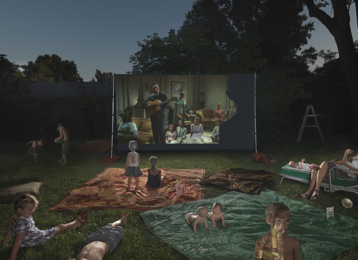 A backyard scene at dusk showing families gathered on blankets and loungers on the grass, watching a projected film on a large outdoor screen. Children play in the foreground while adults relax nearby, with illuminated trees silhouetted against the twilight sky framing the domestic, nostalgic tableau.