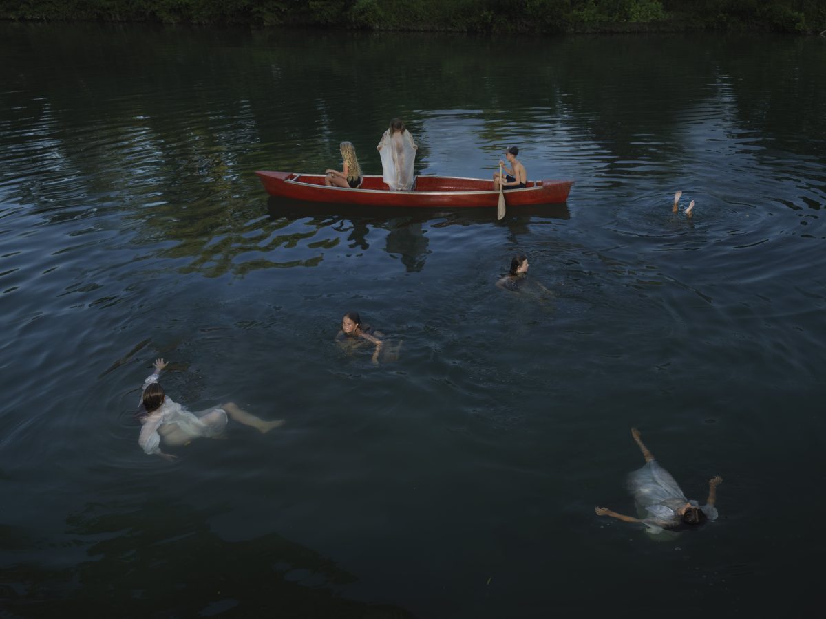 Multiple figures in white clothing swim in dark water around a red rowboat, with two people seated in the boat and dense green foliage visible along the shoreline in the background.