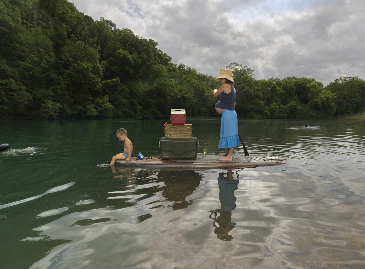 A woman in a straw hat and blue skirt stands on a paddleboard in shallow river water, holding a paddle and gazing outward. A young child sits in the water beside her, while various domestic items—a cooler, picnic basket, and other supplies—are stacked on the board behind her. Dense green forest lines the riverbank under an overcast sky.