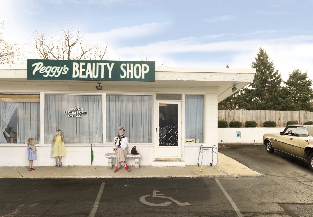 A cream-colored storefront with a green sign reading "Peggy's Beauty Shop" features large windows with white curtains. A woman sits on a bench outside with shopping bags while two young girls in pastel dresses stand nearby; a vintage yellow convertible is parked to the right, and bare trees and evergreens are visible against a pale blue sky.