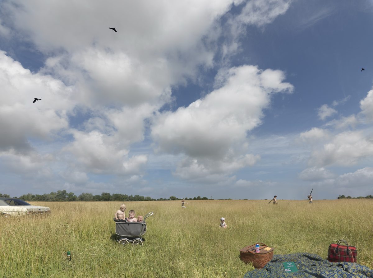 An expansive golden grassland under a blue sky dotted with white clouds. In the foreground, picnic supplies including a wicker basket and plaid blanket rest on the grass. An adult pushes a pram containing a child deeper into the field, while several other figures scatter across the meadow engaged in leisure activities. Birds fly overhead, and distant trees line the horizon.