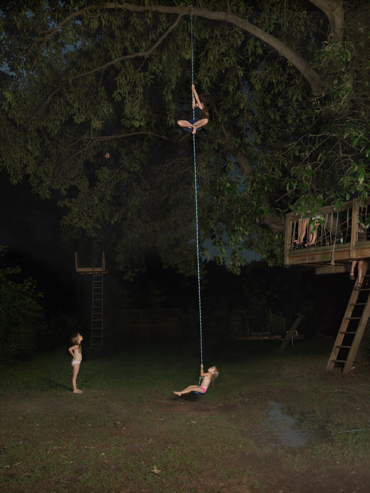 Three shirtless children play on a rope swing suspended from a large tree branch in a nighttime backyard scene. One child hangs from the rope while two others stand on the grass below, illuminated against the dark foliage and a wooden structure visible in the background.