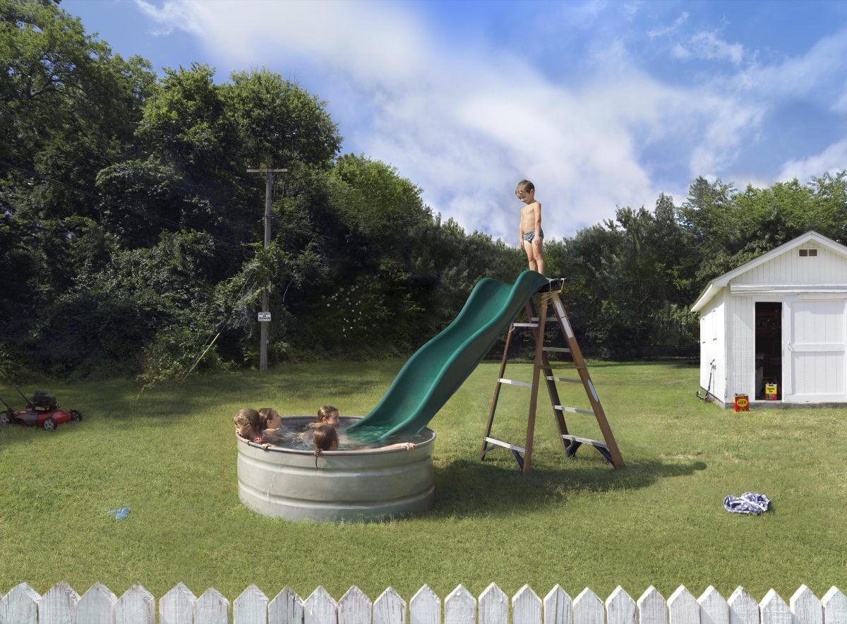 A child stands atop a green slide attached to a wooden ladder frame positioned over a large metal stock tank filled with water, where three other children play. The scene is set in a suburban backyard with manicured lawn, a white storage shed, utility pole, tree line, and white picket fence in the foreground.