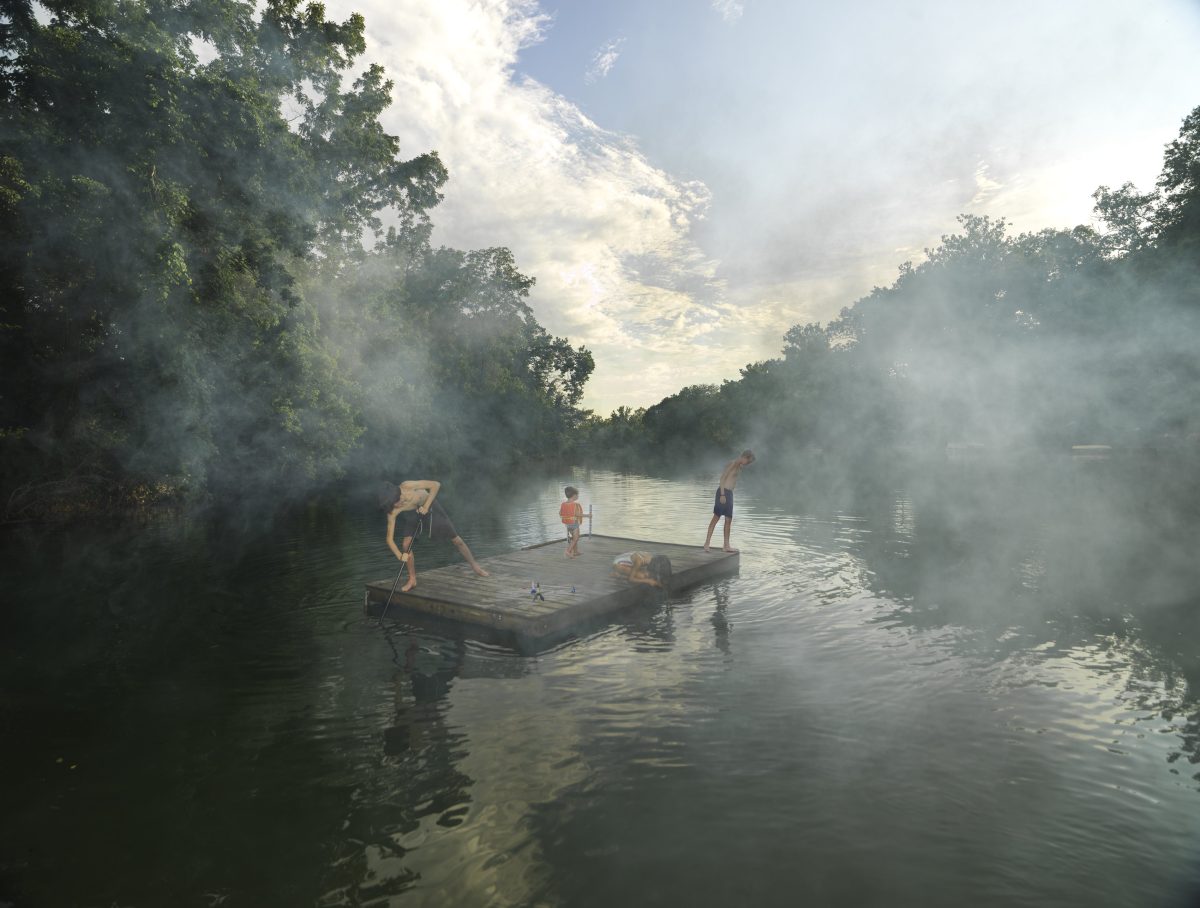 Children and adults play on a wooden raft in a misty river surrounded by dense green forest, with dramatic clouds and light breaking through the canopy above, creating an atmospheric scene of leisure and exploration in nature.