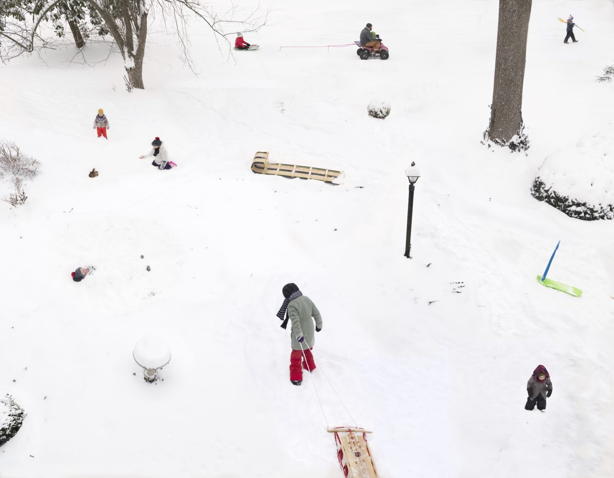 Aerial view of a snowy hillside with multiple people engaged in winter activities scattered across the landscape, including sledding, snowmobiling, and walking. The composition captures the scene from above, with bare trees, evergreen shrubs, and various wooden sleds distributed throughout the white expanse, creating a lively tableau of winter recreation.