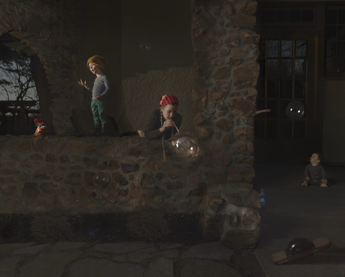 Three children play in a shadowed stone courtyard lit by warm, directional sunlight. One child in a yellow hat stands against the wall on the left, another in a red bandana blows bubbles in the center, and a third sits in darkness on the right. The weathered stone walls and sparse setting create a contemplative atmosphere punctuated by the ephemeral quality of soap bubbles.