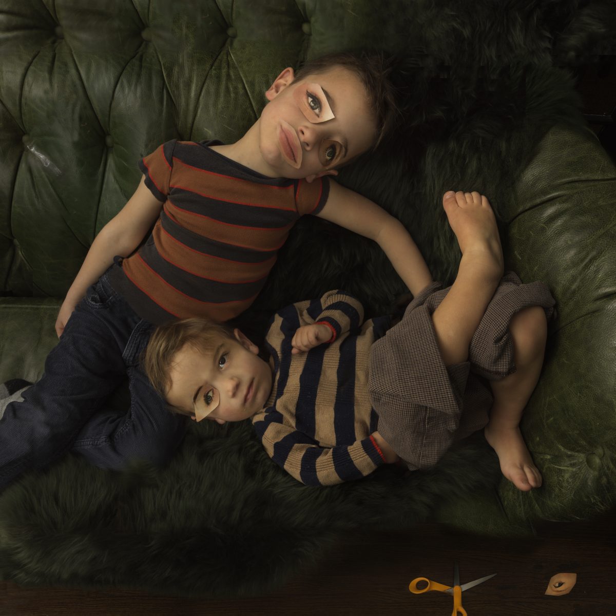 Two young boys lie on a green quilted surface viewed from above, wearing striped shirts in rust and navy tones. The older child gazes directly at the camera while cradling the younger boy, who looks upward with a contemplative expression. A pair of yellow scissors and small objects are visible at the bottom edge of the composition.