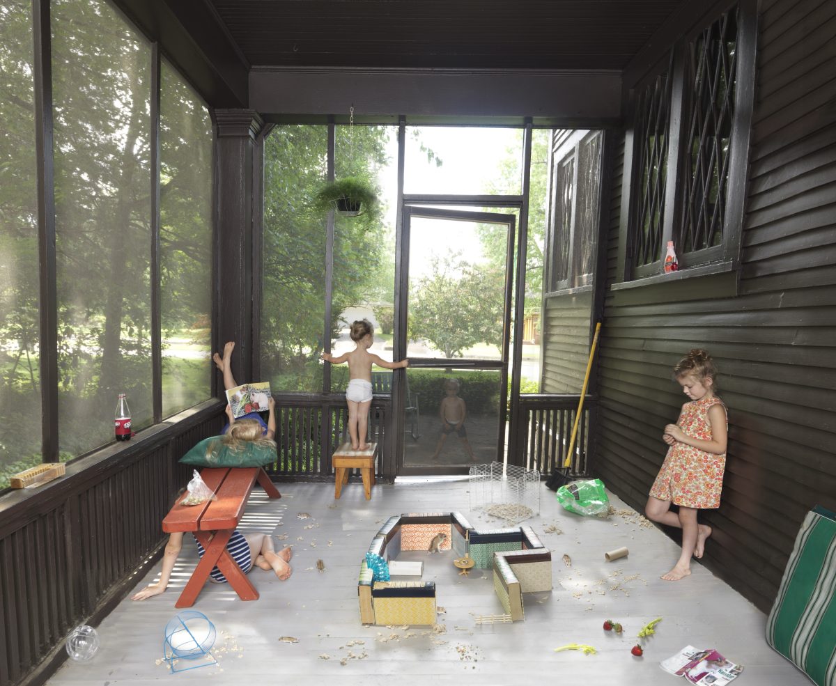 Three young children play in a screened porch surrounded by large windows overlooking green foliage. The concrete floor is scattered with toys, sand, and various containers, while furniture including a red bench and wooden stool are positioned throughout the sunlit space. The composition captures a candid moment of domestic play and exploration in bright, naturalistic tones.