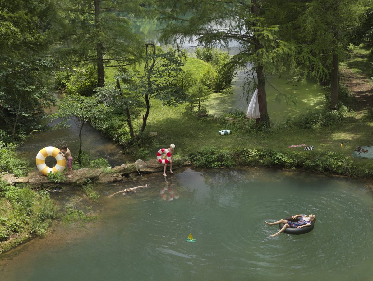 Aerial view of a lush green forest clearing with a winding stream where multiple people enjoy summer leisure activities. A person in a red and white floatie stands at the water's edge, another floats on a tube in the center, while swimmers and sunbathers populate the banks and water, surrounded by dense trees, inflatable toys, and an umbrella.