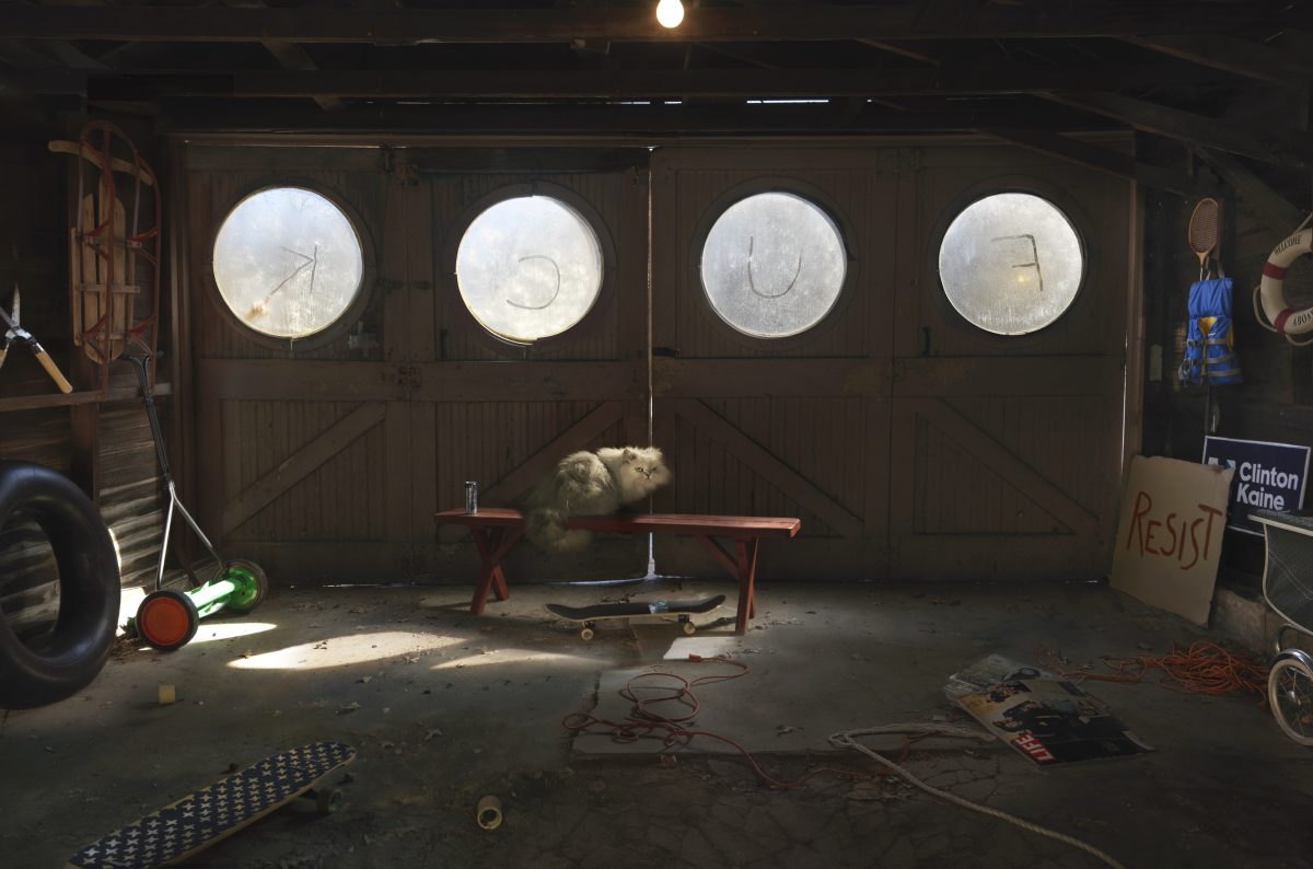 A cluttered garage interior with four large circular windows above wooden doors. Two cats sit on a red bench beneath the windows. Scattered around the concrete floor are various objects including bicycles, a tire, skateboard, tools, electrical cords, and a cardboard sign reading "RESIST" with "Clinton Kaine" visible nearby. Sunlight streams through the circular windows, illuminating the dusty, disorganized space.