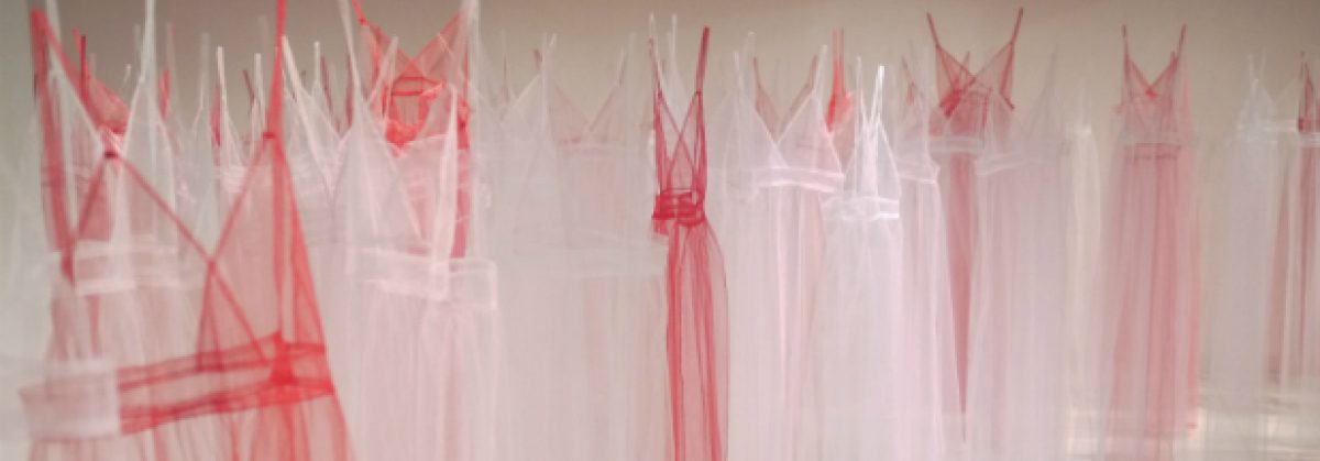 Multiple translucent pink and white tulle garments hang from invisible threads against a neutral wall, their layered, veiled forms creating a composition of transparency and fragility that evokes both intimate domesticity and vulnerability.