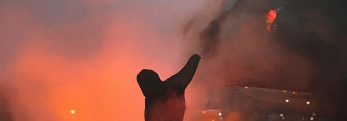 Silhouetted figure with raised arm against a dramatic backdrop of orange and red flames and smoke, rendered in oil painting with gestural brushwork that emphasizes the intensity of the scene.