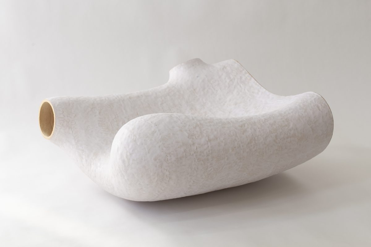White ceramic vessel with organic, undulating forms resembling natural geological shapes. The sculptural object features a hollow cylindrical opening on one side and sits on a neutral background, displaying the subtle texture and matte finish characteristic of hand-built clay work.