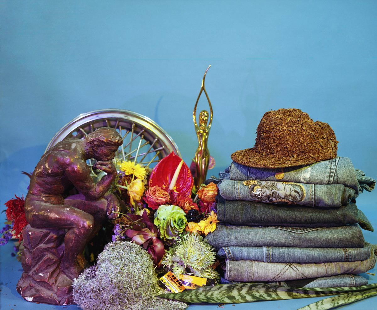 A vibrant still life arrangement against a blue background featuring a purple sculptural figure on the left interacting with a Ferris wheel and colorful flowers, a golden statue in the center, and a stack of folded textiles topped with a cork hat on the right, combining found objects, natural elements, and symbolic imagery.
