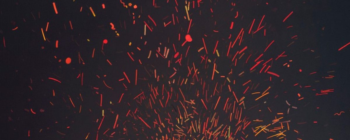 Abstract composition of dynamic red and orange streaks and particles radiating outward against a dark background, creating an energetic burst effect suggestive of fire, sparks, or explosive movement.