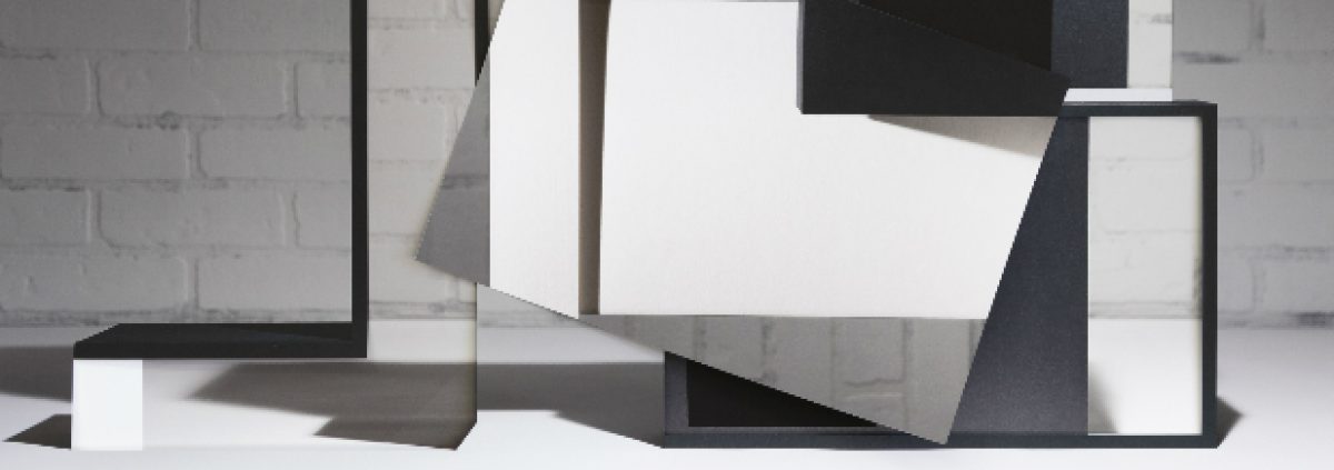 Abstract geometric sculptural installation featuring white and dark forms arranged on a white platform against a brick wall, with angular planes and rectangular elements creating a composition that explores spatial relationships and modernist architectural principles.