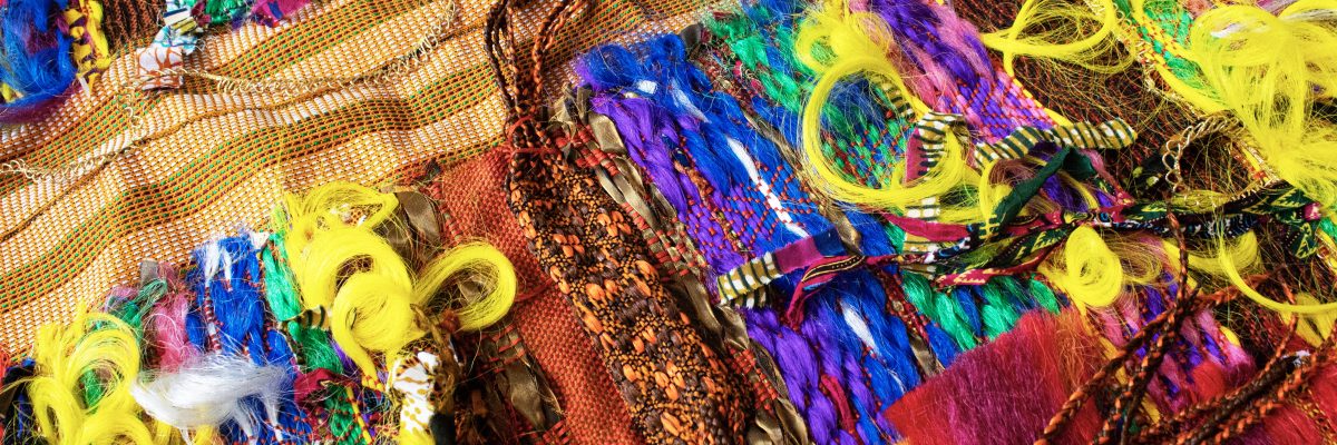 Vibrant textile artwork featuring intricately woven and braided synthetic hair in bold colors including yellow, blue, purple, pink, and orange, layered over patterned fabrics with geometric designs and mesh textures, creating a dense, three-dimensional composition that celebrates African diaspora aesthetics and hair culture.