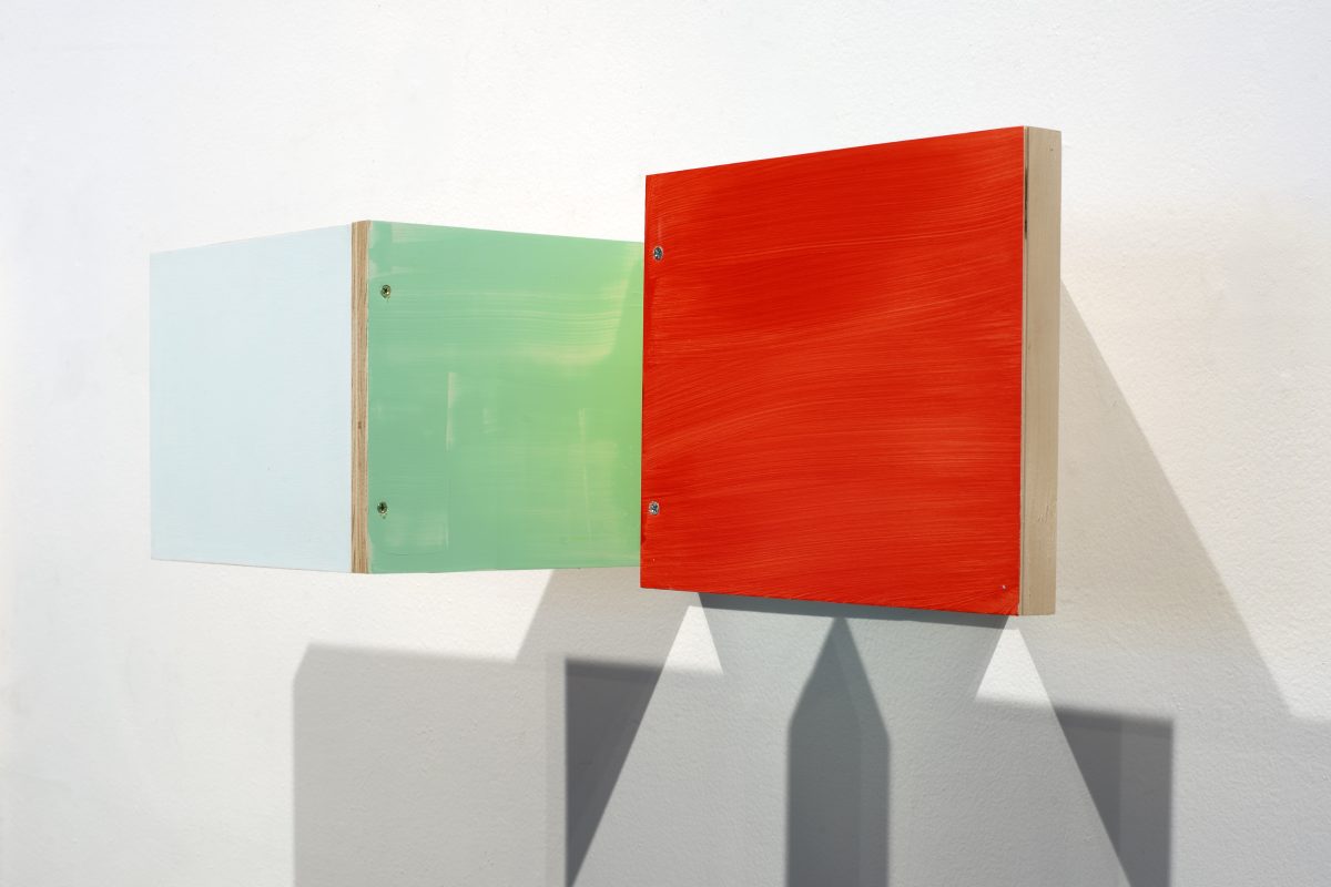 Three wall-mounted boxes with natural wood edges and colored front panels—pale blue, soft green, and vibrant red—arranged in a horizontal line, casting geometric shadows on a white wall.