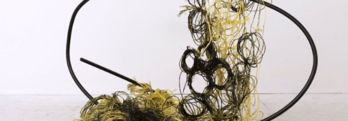 A sculptural installation featuring tangled coils of golden-yellow and black materials suspended within and around a curved black steel frame, creating a dynamic composition that suggests both containment and overflow against a white gallery wall.
