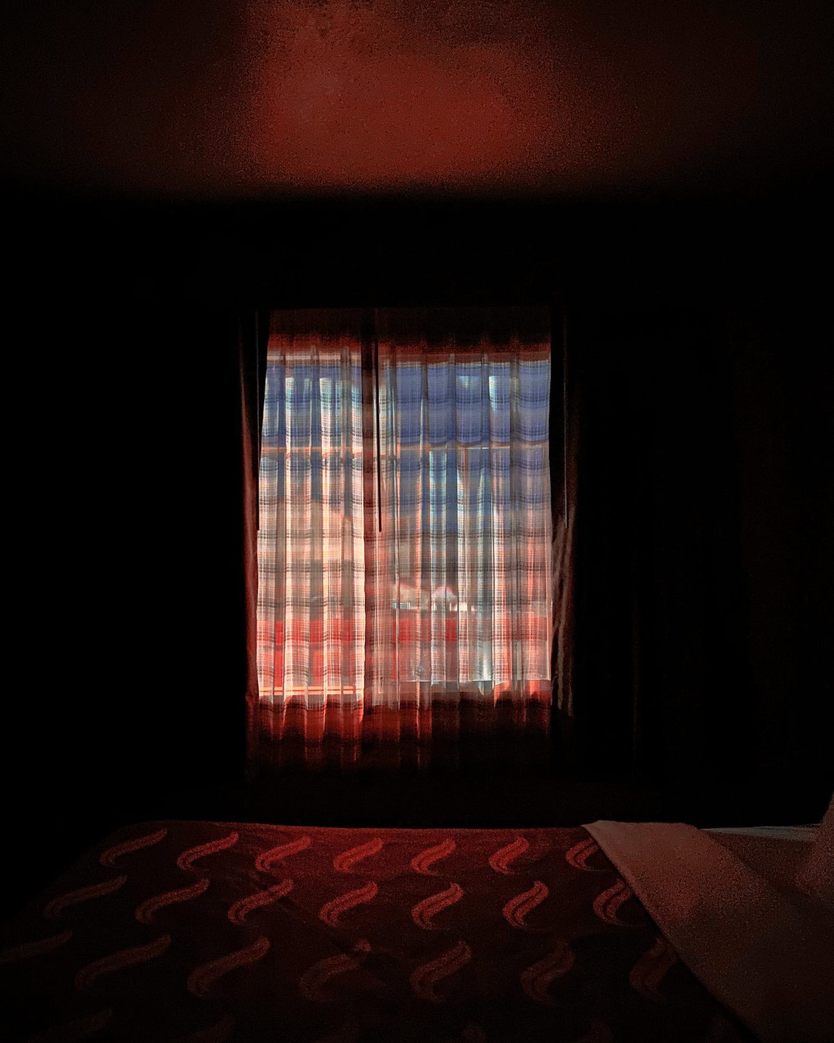 A darkened interior room with a window framed by red curtains that glow warmly in the filtered light. The window reveals a distant cityscape beyond sheer curtains, rendered in cool blues and oranges. In the foreground, a patterned red textile with geometric motifs occupies the lower portion of the composition, creating a strong contrast between the intimate domestic space and the distant urban landscape visible through the glass.