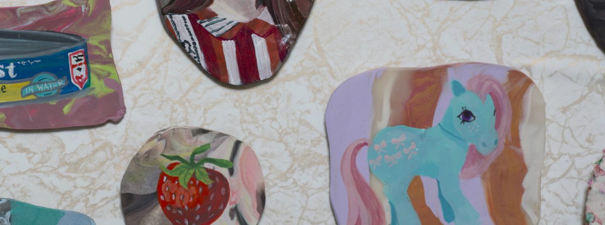 Scattered circular and curved artworks on a cream-colored textured surface featuring vibrant imagery including a turquoise unicorn with pink mane, a red strawberry, red and white striped patterns, and packaged items, combining painting with decorative domestic references in a mixed-media arrangement.