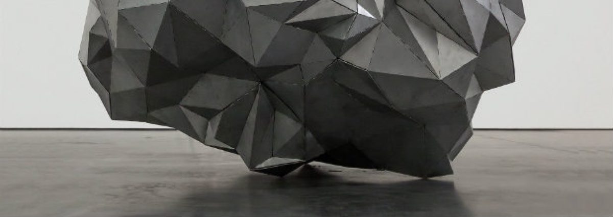 Large geometric sculpture composed of angular, faceted planes in shades of gray and black, positioned on a concrete floor against a white wall. The form creates a complex, crystalline volume with sharp edges and overlapping surfaces that suggest both architectural and organic qualities.