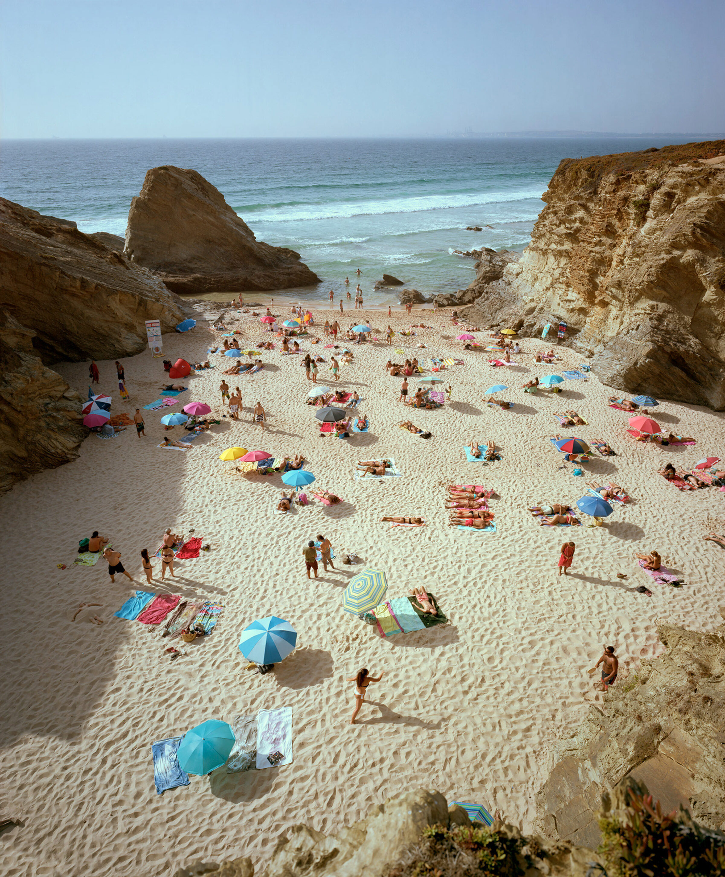 Aerial view of a crowded beach nestled between towering ochre-colored cliff formations, with numerous beachgoers scattered across pale sand beneath colorful umbrellas in shades of blue, pink, and yellow. Turquoise waves and rocky outcroppings frame the scene, while the vast horizon extends across a clear sky.