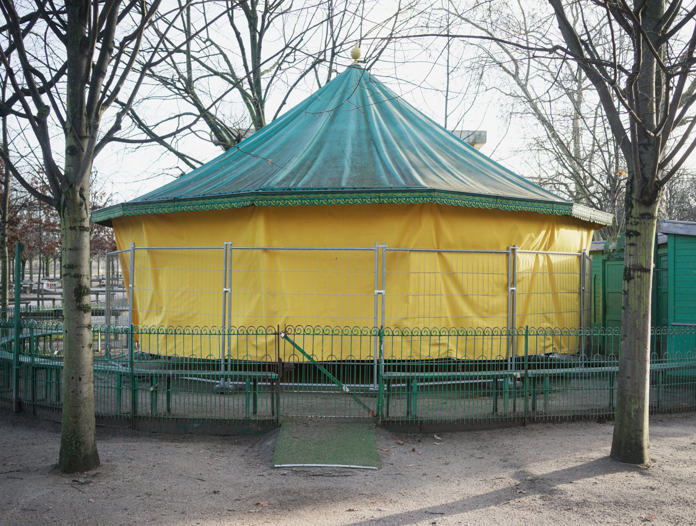 A circular bandstand with a turquoise conical roof and yellow fabric walls stands in a park, framed by bare trees and surrounded by green metal fencing. The structure sits on a gravel ground with ornamental railings, creating a striking contrast of bright colors against the muted winter landscape.