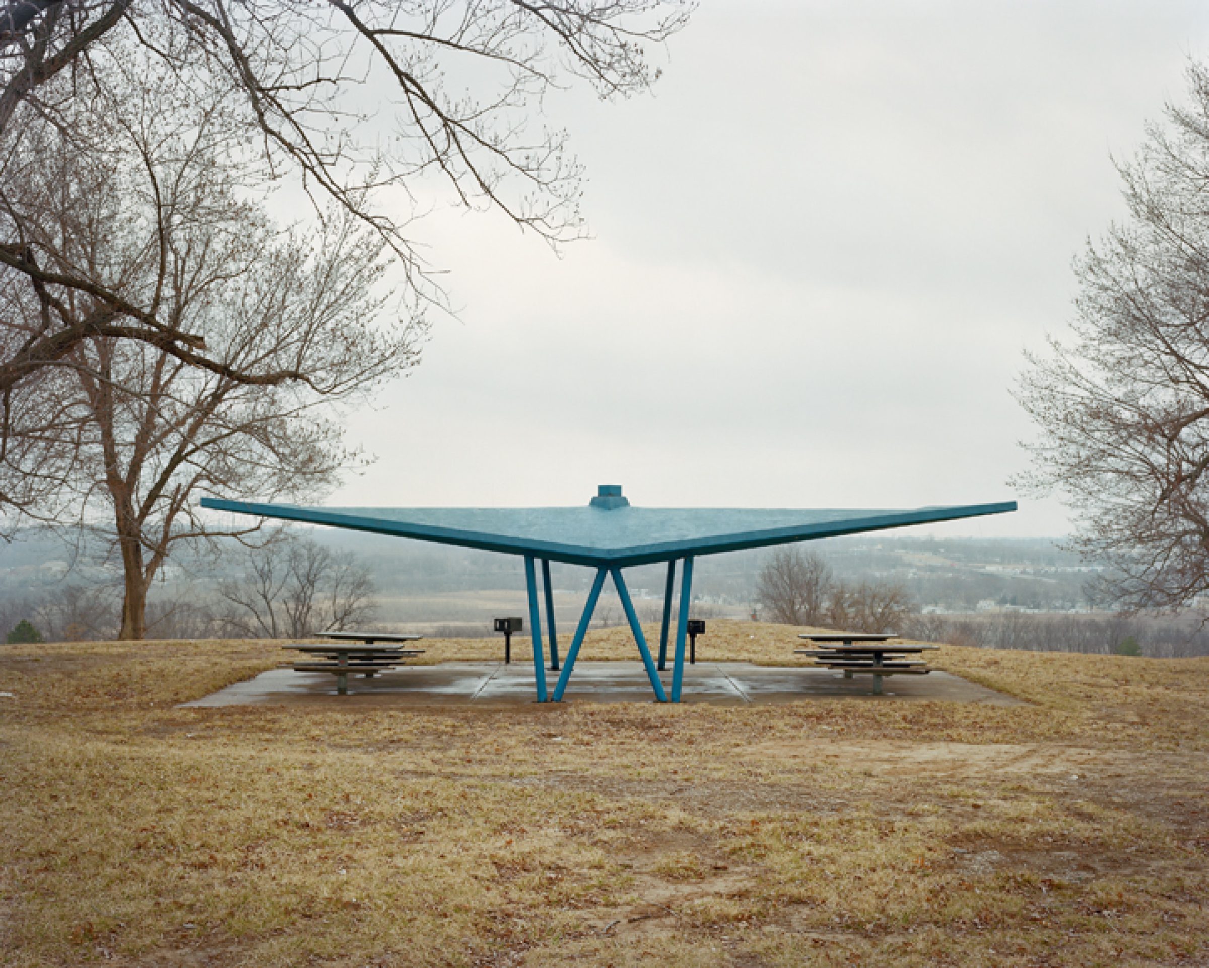 A large teal-blue sculptural shelter with an expansive wing-like roof structure stands in a sparse winter landscape. Bare trees frame the composition on either side, with picnic tables positioned beneath the canopy and rolling hills visible in the distant background.