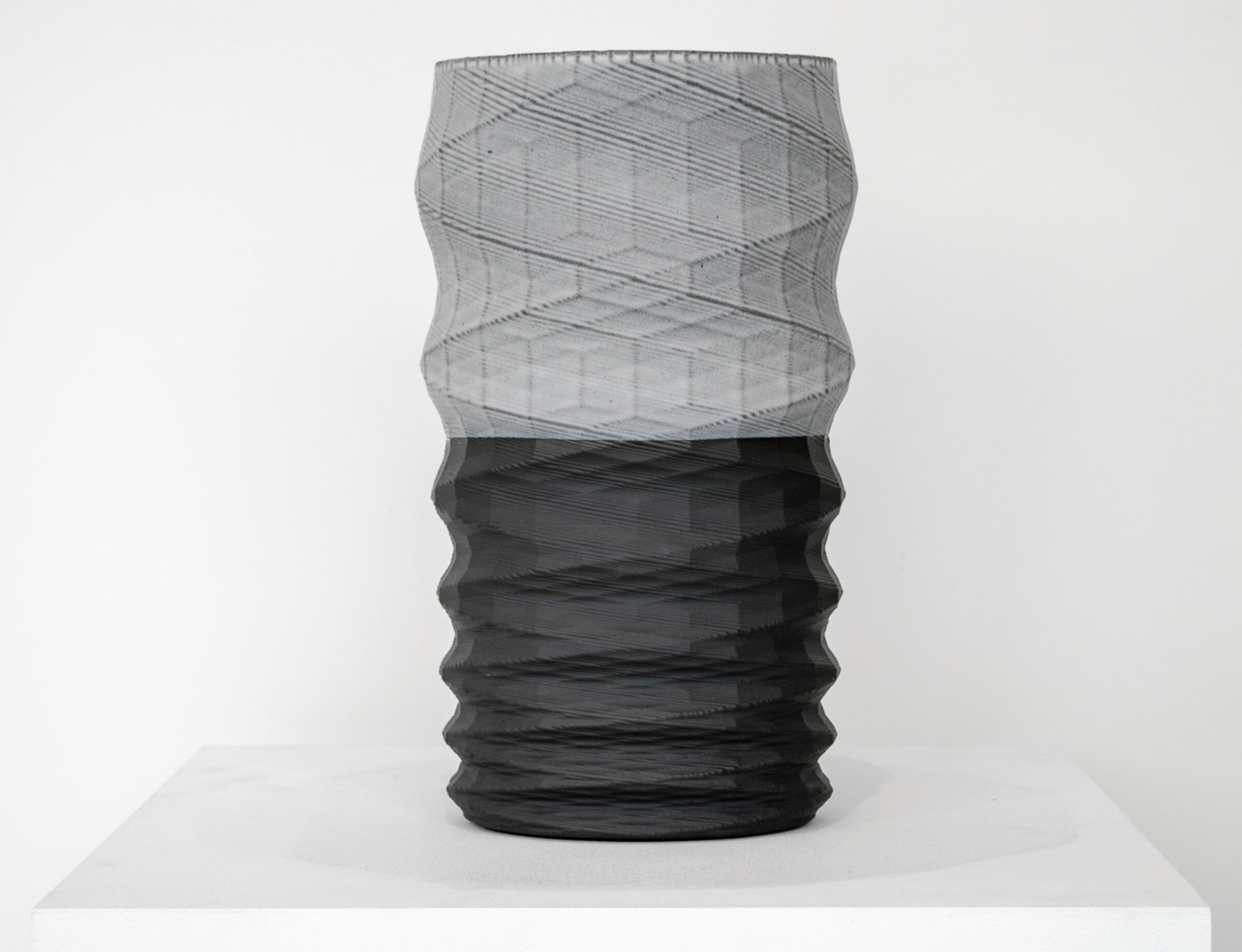 A tall cylindrical vessel with a twisted, ribbed surface sits on a white platform. The upper half features a translucent gray mesh-like texture, while the lower half is rendered in solid black with horizontal ridging, creating a striking color and material contrast.
