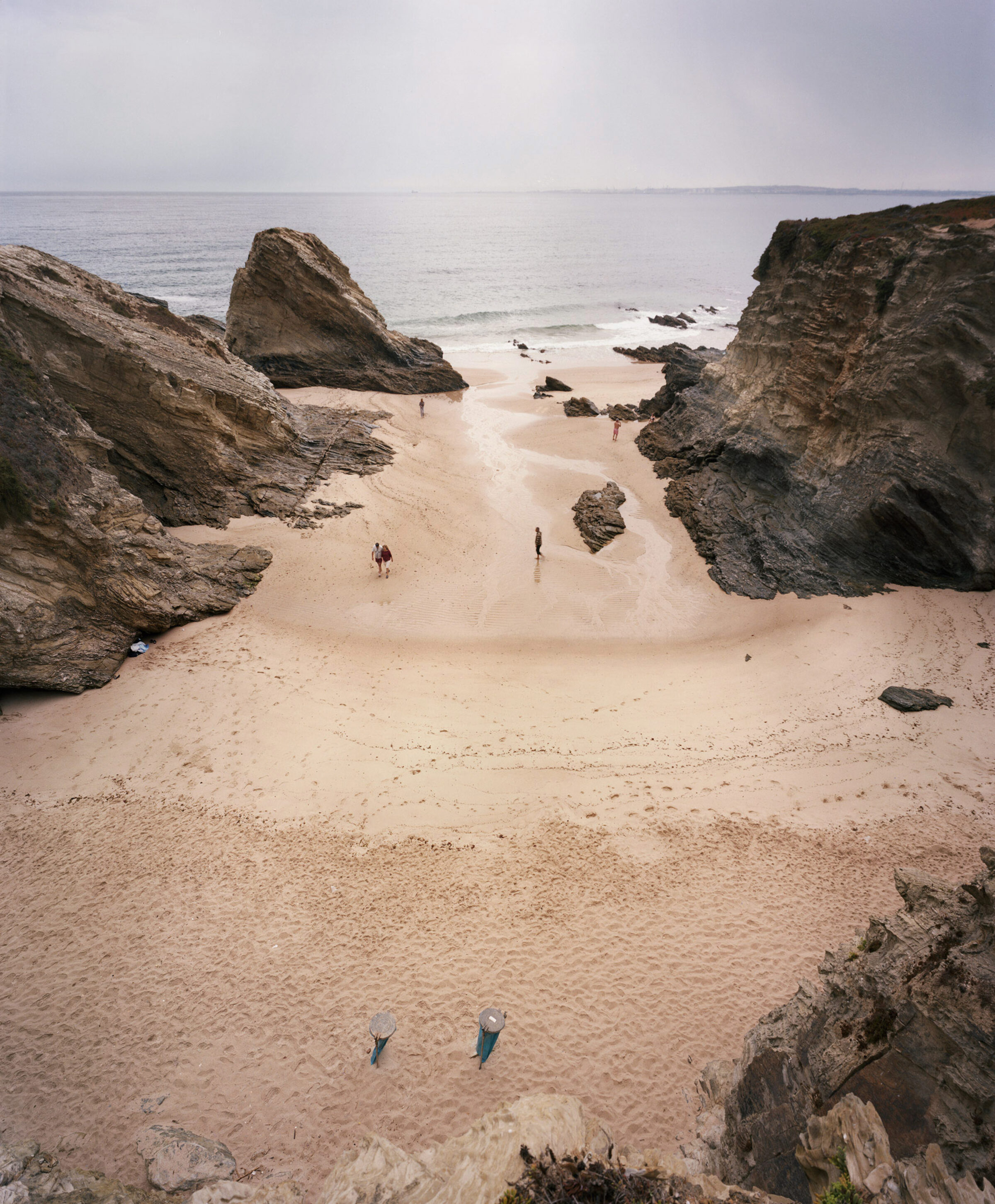 A secluded sandy beach enclosed by dramatic rocky outcrops and large boulders, with several small figures scattered across the sand and wading in shallow water. The composition is framed by weathered stone formations in the foreground and middle ground, while calm ocean and pale sky occupy the background.