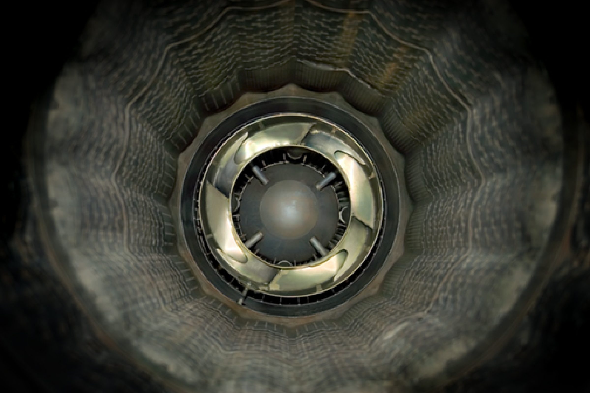 A symmetrical view down a cylindrical tunnel with concentric ribbed walls in gray and black tones, centered on a metallic circular aperture with reflective surfaces and a dark spherical core.