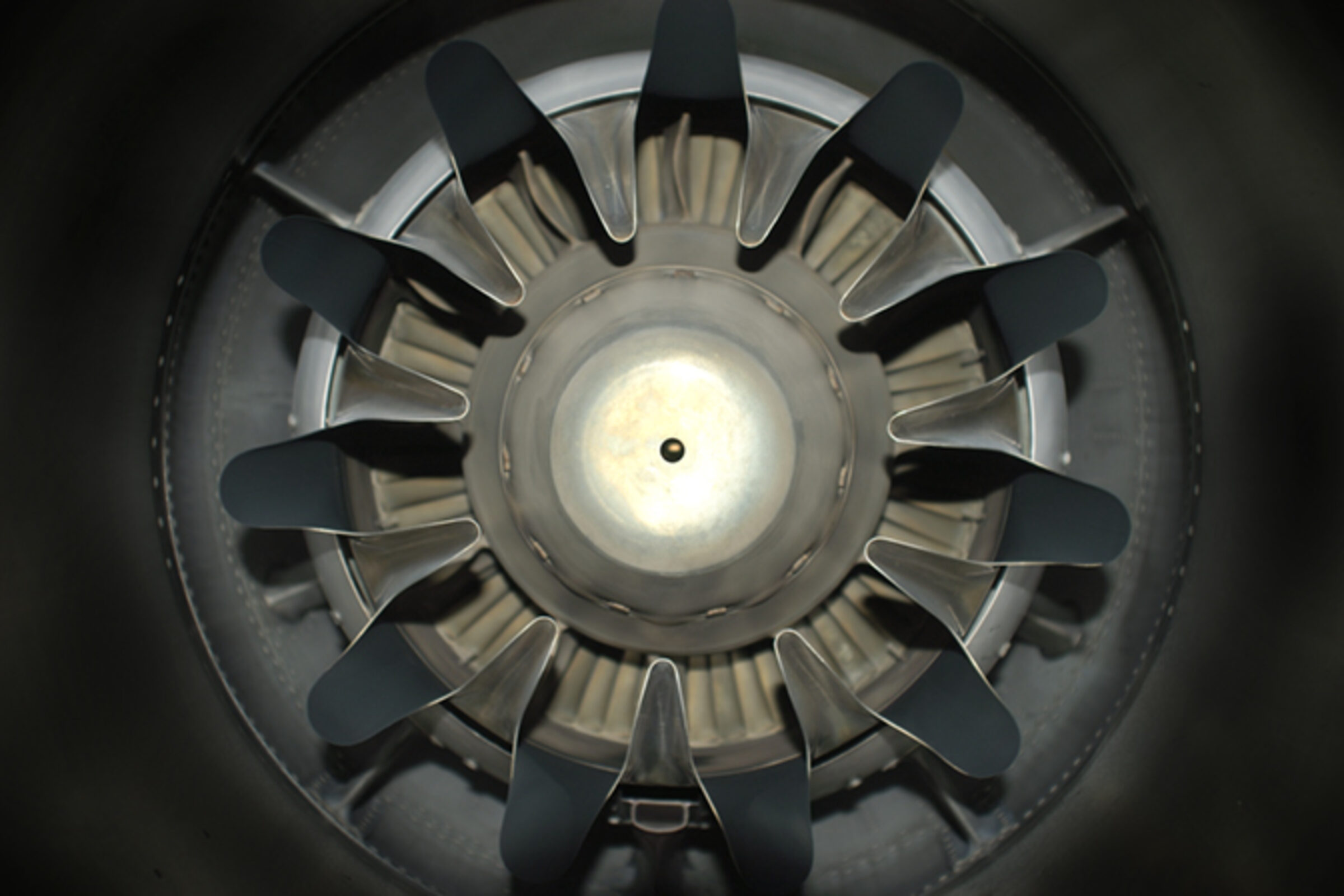 Overhead view of a circular mechanical structure with black blade-like forms radiating outward from a pale central hub, photographed against a dark background. The composition emphasizes radial symmetry and industrial materiality, with metallic and matte surfaces creating subtle tonal contrasts.