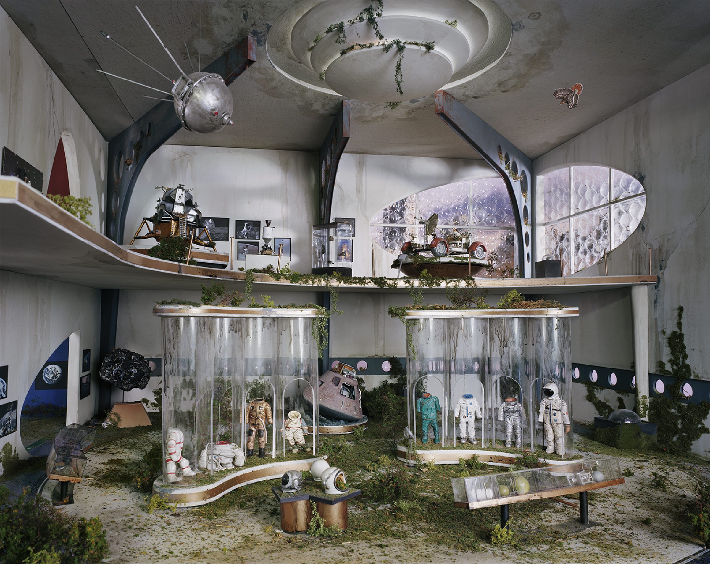 An abandoned institutional interior overgrown with vegetation features glass display cases containing astronaut suits and space equipment, with a Soyuz capsule and lunar rover visible on upper shelves. Concrete walls, debris-strewn floors, and celestial imagery suggest a decaying space museum frozen in time.