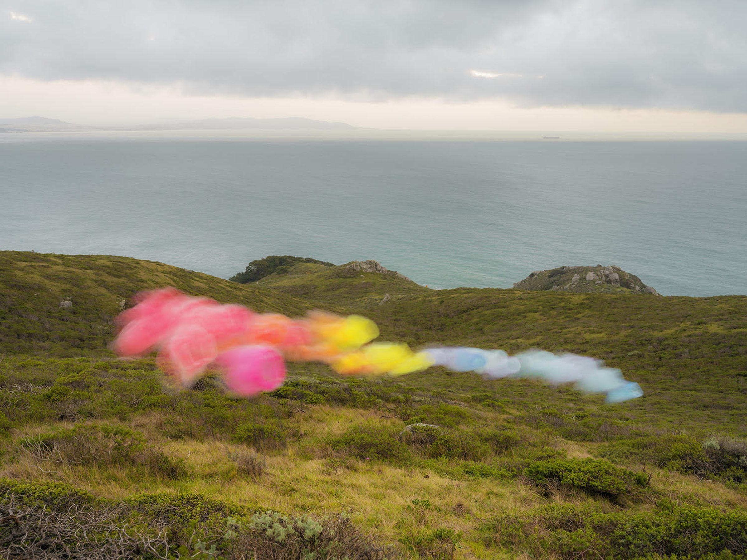 Blurred rainbow-colored tulle fabric streaks across a green coastal hillside overlooking turquoise ocean waters under an overcast sky, creating an ethereal contrast between the delicate, translucent material and the rugged landscape.