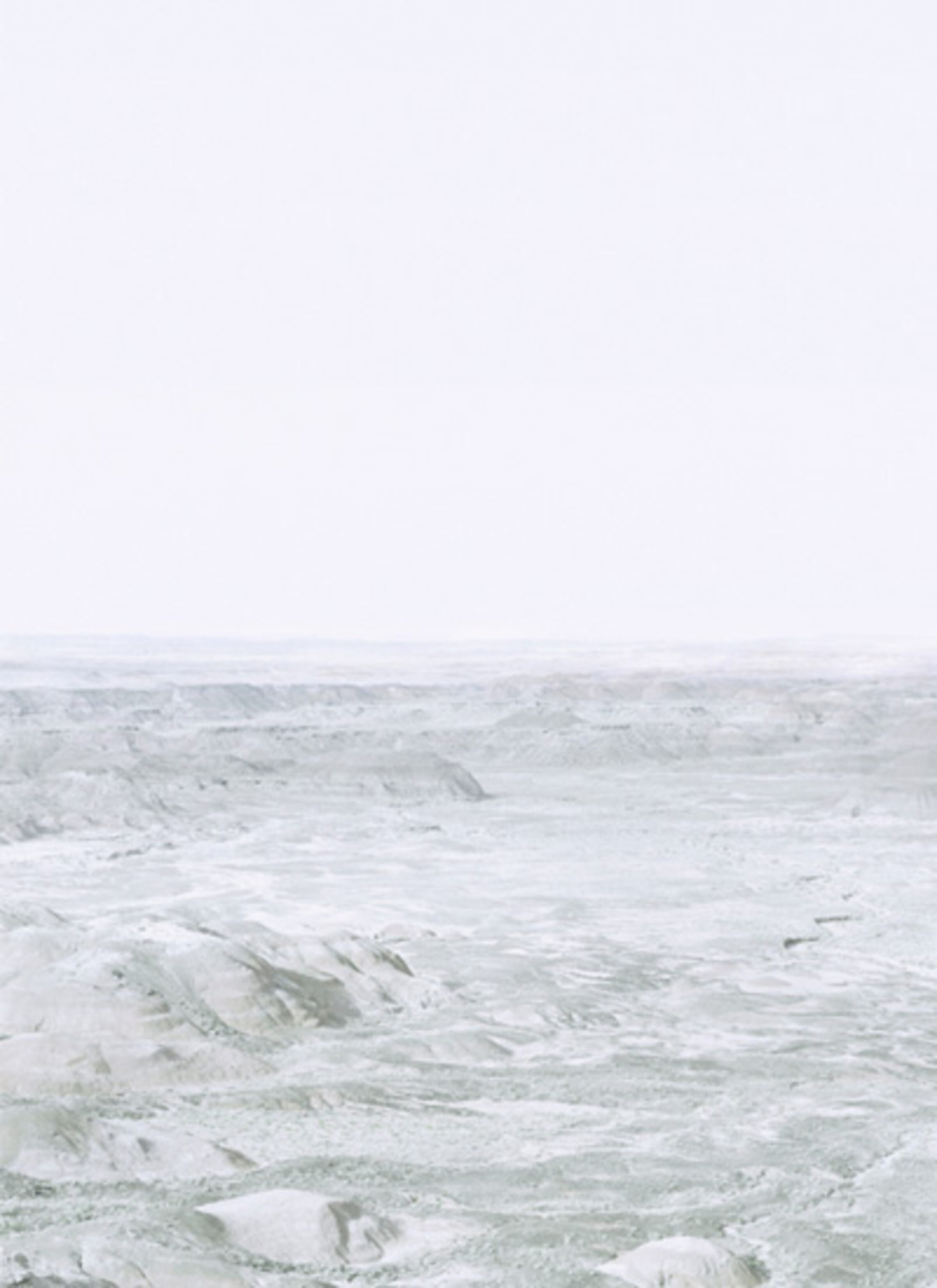 Ethereal landscape photograph of an expansive, barren terrain with undulating hills rendered in soft whites, grays, and pale greens beneath a hazy, overexposed sky. The composition creates an abstracted, almost monochromatic vista that blurs the distinction between earth and atmosphere.