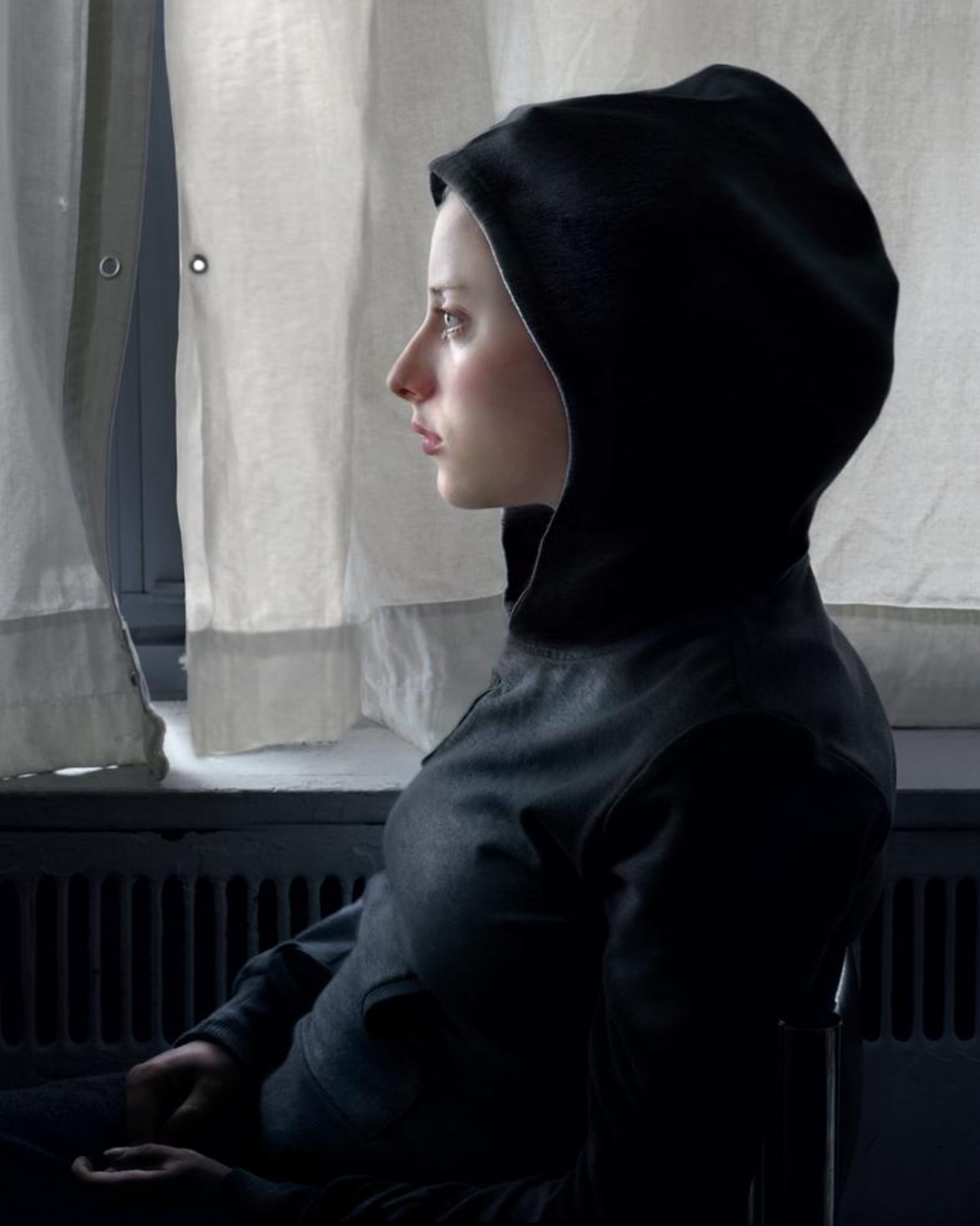 A figure in profile wearing a black hooded garment sits by a window with sheer white curtains. The minimalist composition features soft natural light against neutral tones and a dark radiator below the window sill.