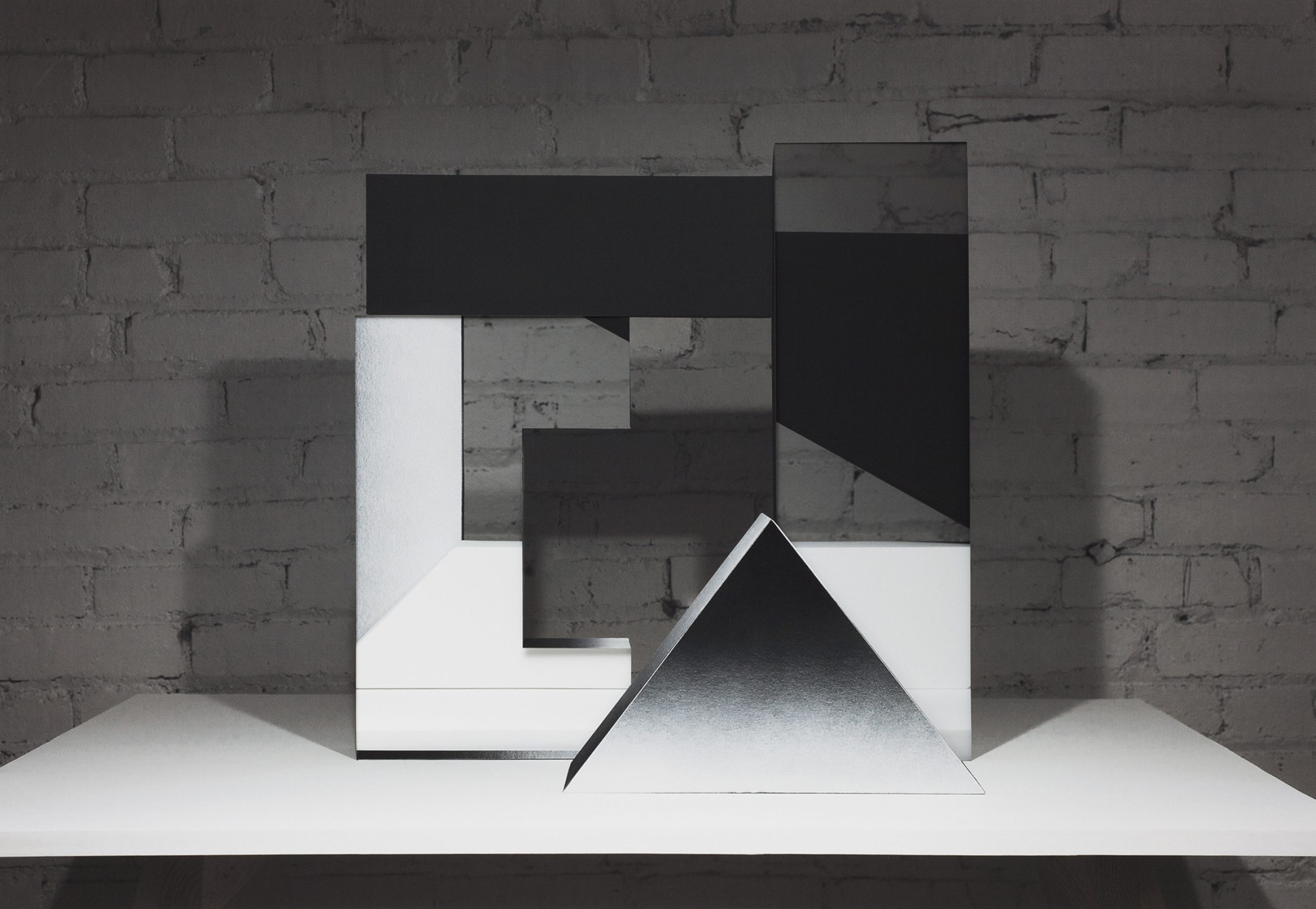 Abstract geometric composition featuring layered black and white rectangular forms arranged in an interlocking pattern against a weathered brick wall. A white triangular prism sits prominently in the foreground on a white platform, casting shadows that emphasize its three-dimensional form.