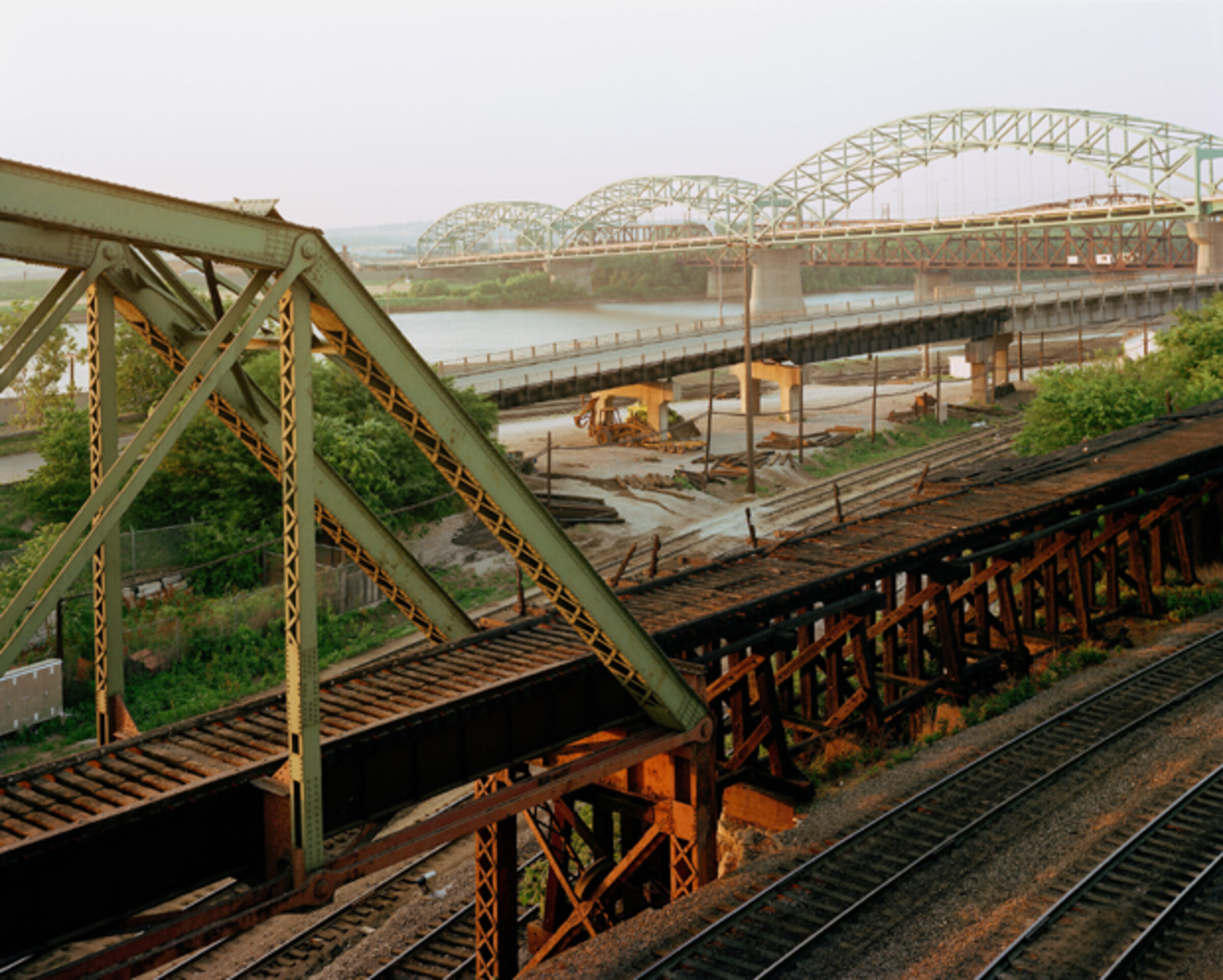 Industrial railroad bridge infrastructure in the foreground with rusted steel trusses and support beams, extending toward a modern arched bridge crossing a wide river in the distance. The composition layers deteriorating metalwork, active rail yards with equipment and structures, and waterway landscape under overcast sky, rendered in muted greens, browns, and grays.